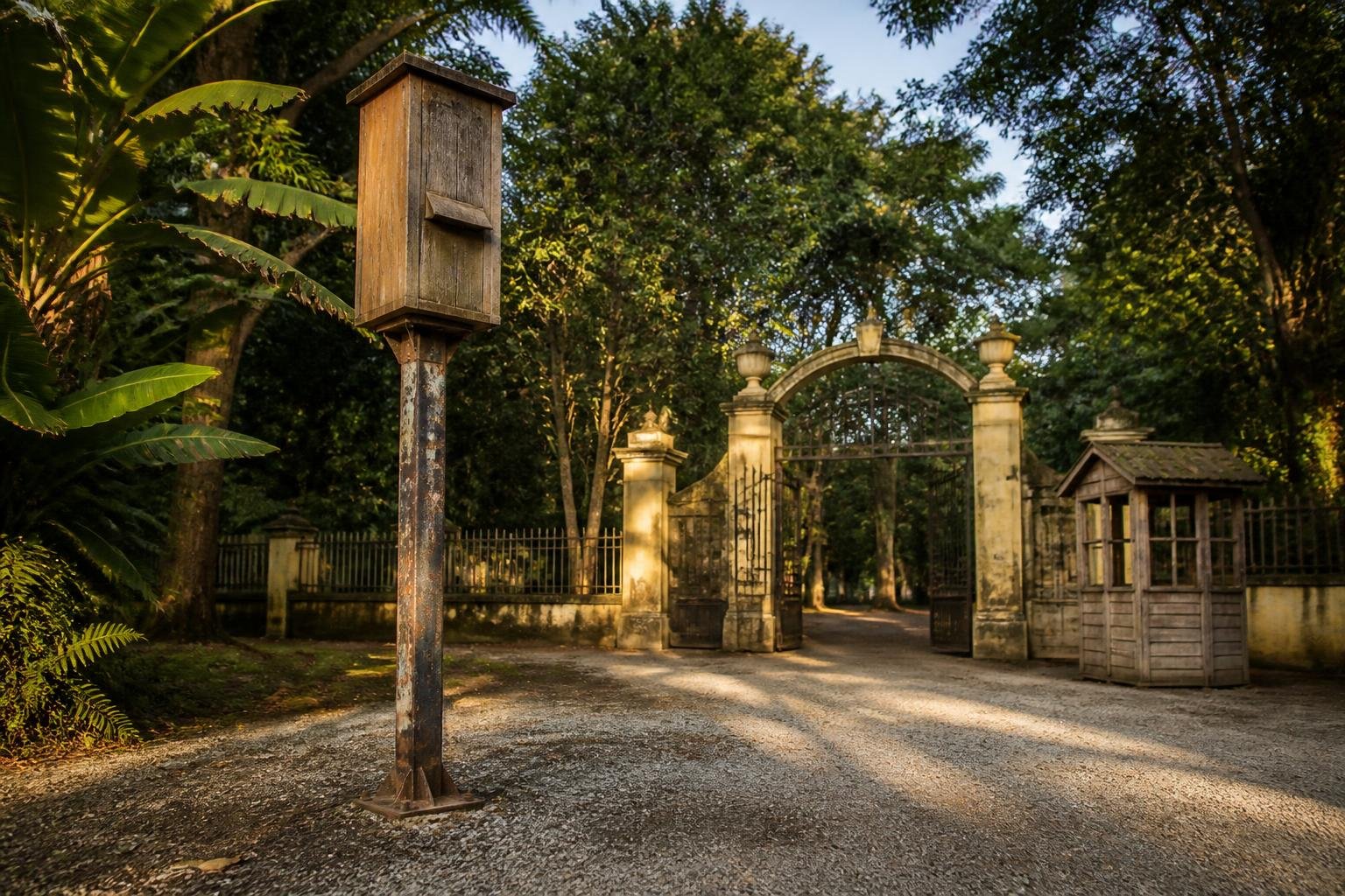 Caixa de madeira suspensa num poste de ferro na entrada do antigo zoológico de Vila Isabel, cercada de vegetação tropical